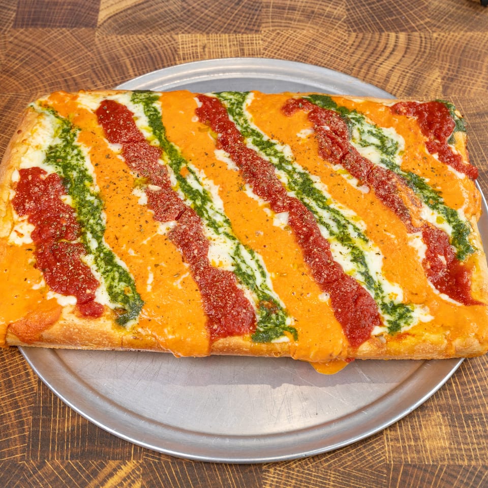 Rectangular pizza with stripes of red sauce, orange sauce, and green pesto on a metal tray.