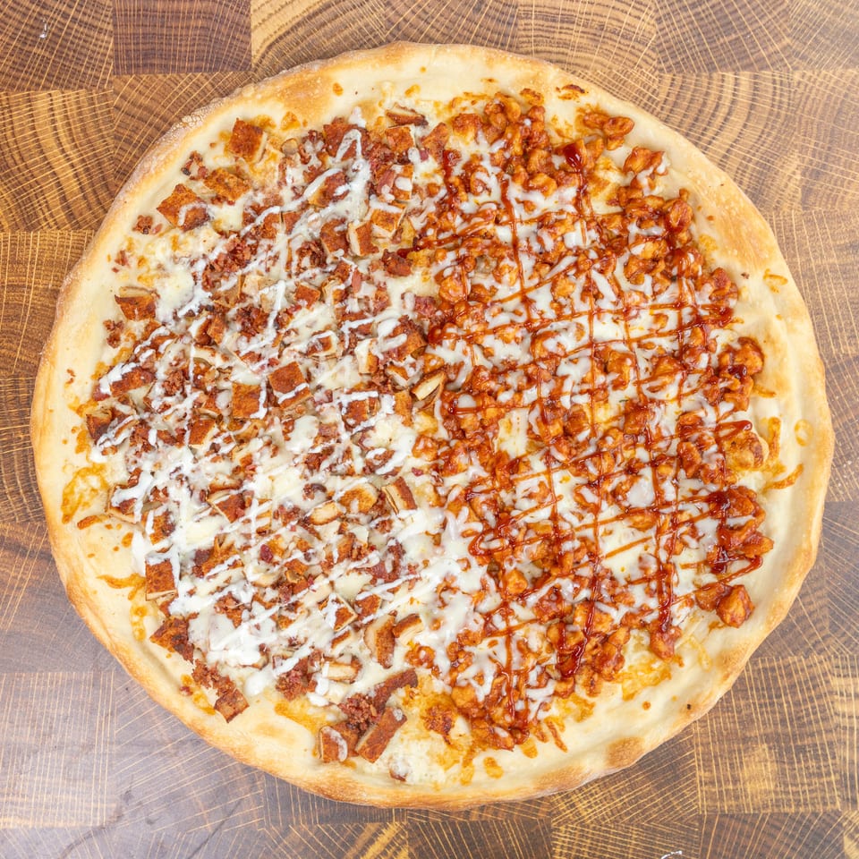 A whole pizza, half with white sauce and bacon, half with red sauce and chicken, on wood.