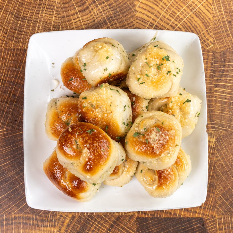 A white square plate filled with golden-brown garlic knots, sprinkled with cheese and parsley, on a wooden table.