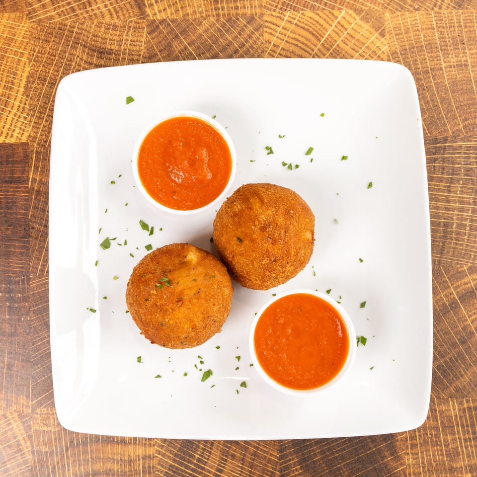 Two golden-brown arancini balls with two small bowls of red marinara sauce on a white plate.